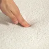 A hand is pressing into a soft, cream-colored textured surface, likely fabric or carpet. Produktbild-Vorschau