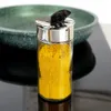 A spice jar filled with yellow spice, placed on a reflective black surface with a blurred green bowl in the background. Produktbild-Vorschau