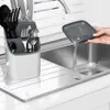 A hand is pouring water from a small tray into a kitchen sink, next to a utensil holder filled with cutlery. Produktbild-Vorschau