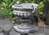 A stone urn is placed on rocks in a garden setting with green foliage in the background. Produktbild-Vorschau