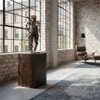 A steampunk-style sculpture of a person holding a rifle, displayed on a wooden pedestal in a loft-style room with large windows and exposed brick walls. Náhled obrázku produktu