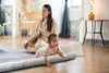 A mother and baby are on a yoga mat in a room. The baby is crawling on a gray bolster pillow. Produktbild-Vorschau