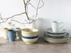 A collection of ceramic dishes and mugs arranged on a wooden surface, with a decorative branch in a pitcher in the background. Náhľad obrázku produktu