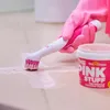 A hand wearing a pink glove is using an electric cleaning brush with pink bristles to scrub a light-colored tiled surface, with a tub of "The Pink Stuff" cleaning paste nearby. Produktbild-Vorschau