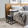 A black side table with a magazine rack is placed next to a bed in a bedroom. Náhľad obrázku produktu