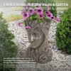 A decorative plant pot shaped like a woman's head, filled with purple petunias, is placed outdoors on a bed of gravel, surrounded by greenery. Náhled obrázku produktu