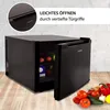 A black mini-fridge with its door ajar, showing bottles and a carton inside. The fridge is on a wooden surface with some vegetables in the foreground. The background is a textured wall. Produktbild-Vorschau