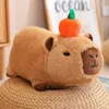 A plush toy capybara with a small orange and green apple on its head, sitting on a woven stool. Náhled obrázku produktu