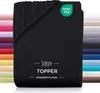 A black jersey topper fitted sheet is shown folded, with a stack of other colored fitted sheets visible behind it. Produktbild-Vorschau