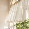 A close-up view of a cream-colored lace curtain with tassels, hanging in a window with sunlight streaming through. Green foliage is visible outside the window. Produktbild-Vorschau