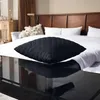 A black quilted pillow is placed on a reflective black coffee table in front of a bed with white bedding and a wooden headboard. Produktbild-Vorschau