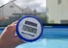 A hand holds a thermometer showing 29.9 degrees Celsius in front of a swimming pool. Produktbild-Vorschau