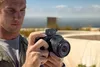 A man is holding a camera with a microphone attached, outdoors with a blurred background of water and a railing. Voir les images du produit