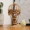 A wooden 3D model of a hot air balloon sits on a wooden surface, with a potted plant and a white rose in the background. Produktbild-Vorschau