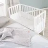 A white wooden crib with a mattress and bedding, placed next to a larger bed. Produktbild-Vorschau