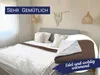 A bedroom scene with a bed made up with a white fluffy blanket and a brown blanket folded on top. Decorative elements like macrame and woven lampshades are visible. Produktbild-Vorschau