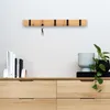 A wooden key rack with black accents is mounted on a white wall above a wooden dresser. A set of keys hangs from one of the hooks. On the dresser, there are books, a decorative pot, and a plant. Produktbild-Vorschau