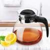 A glass teapot filled with amber liquid, with lemon slices and leaves on a white wooden surface. Produktbild-Vorschau
