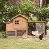 A wooden chicken coop with an attached run is set up in a grassy yard with lush green foliage and a brick building in the background. Produktbild-Vorschau