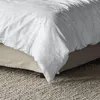 A close-up view of a duvet cover on a bed, showing the texture and drape of the fabric. The bed is covered with a beige bed skirt, and a textured rug is visible on the floor. Produktbild-Vorschau