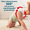 A baby is doing a handstand on a carpeted floor, wearing a diaper. Red arrows indicate a 360-degree fit around the diaper. Podgląd zdjęcia produktu