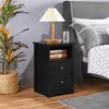 A black nightstand with two drawers and an open shelf, placed next to a bed in a bedroom setting. A lamp, books, and a glass of water are on top of the nightstand. A large potted plant is to the left. Podgląd zdjęcia produktu