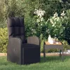 A black rattan garden recliner chair with a cushion and a small side table with a bottle and a bowl of fruit, placed on a green lawn with lush green foliage and white flowers in the background. Produktbild-Vorschau