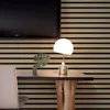 Goldene und weiße pilzförmige Tischlampe auf einem dunklen Holzschreibtisch mit Pflanze, vor einer Lamellenholzwand Produktbild-Vorschau