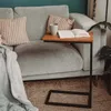 A C-shaped side table with a wooden top is placed next to a sofa, holding a book and a mug. A blanket is draped over the sofa. Produktbild-Vorschau