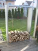 A metal firewood rack filled with logs is placed on a wooden deck in a backyard setting. Produktbild-Vorschau
