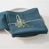 A stack of teal napkins tied with a string and a small white flower on top, placed on a white plate. Produktbild-Vorschau