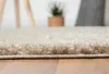 A close-up, low-angle view of a beige shaggy rug on a wooden floor. In the background, out of focus, are parts of furniture and a potted plant. Náhľad obrázku produktu