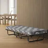 A folding guest bed with a mattress is shown in a room with wooden walls and furniture in the background. Náhľad obrázku produktu