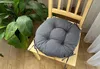 A round gray seat cushion is placed on a light wooden chair next to a large green plant. Produktbild-Vorschau