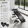 A sit-up bench with dumbbells and resistance bands in a modern indoor setting with large windows. Náhled obrázku produktu