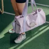 A person holding a purple duffel bag with a Pusheen character on it, standing on a green surface with white lines. Produktbild-Vorschau