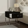 A black high-gloss nightstand with a chrome metal frame is placed next to a bed. Produktbild-Vorschau