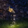 A solar-powered watering can light string is displayed in a garden at night, with a stack of firewood in the background. Produktbild-Vorschau