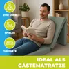 A man is reading a book while sitting on a folded mattress in a living room. Produktbild-Vorschau