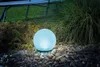 A glowing blue sphere sits on a bed of pebbles in a garden setting, next to lush green grass and plants. Produktbild-Vorschau