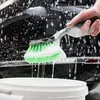 A hand holding a car wash brush with green bristles and white foam, dripping into a white bucket, with a soapy black car in the background. Produktbild-Vorschau