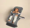 A baby is sitting in a gray Ergobaby bouncer against a plain background. Produktbild-Vorschau