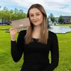 A young woman is holding a beige wallet in a park with a fountain in the background. Produktbild-Vorschau