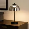 A modern black table lamp with a smoked glass shade is placed on a dark wooden dresser. Produktbild-Vorschau