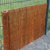 A rolled-up willow privacy screen leaning against a metal fence, with grass and gravel in the background. Produktbild-Vorschau