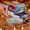 A hand is placing a small model of a starship onto a larger model of a starship, with a gift box and Christmas lights in the background. Podgląd zdjęcia produktu