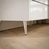 A close-up view of the bottom part of a white dresser with silver legs, resting on a wooden floor. Produktbild-Vorschau