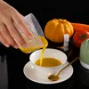 A hand pouring liquid from a measuring cup into a bowl, with vegetables and a mini chopper in the background. Produktbild-Vorschau