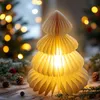 A lit paper Christmas tree decoration sits on a wooden surface, surrounded by pinecones and greenery, with blurred Christmas lights in the background. Produktbild-Vorschau