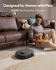 A man is sitting on the floor next to a brown leather sofa, petting a small dog. A robot vacuum cleaner is cleaning the carpet in front of him, and there is a pile of pet hair on the carpet. Voir les images du produit